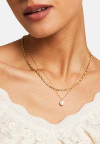 Ogrlice od pozlaćenog metala imaju uvijenu lančić i delikatni lančić s jednim bisernim privjeskom. Postavka je minimalistička i elegantna.