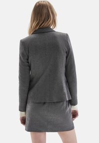 Mujer con cabello rubio largo, vistiendo un blazer texturizado gris oscuro y una falda a juego, vista desde atrás contra un fondo blanco.