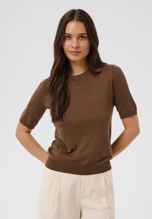 Femme aux longs cheveux foncés portant un haut en maille marron à manches courtes et un pantalon beige clair, debout les mains dans le dos.