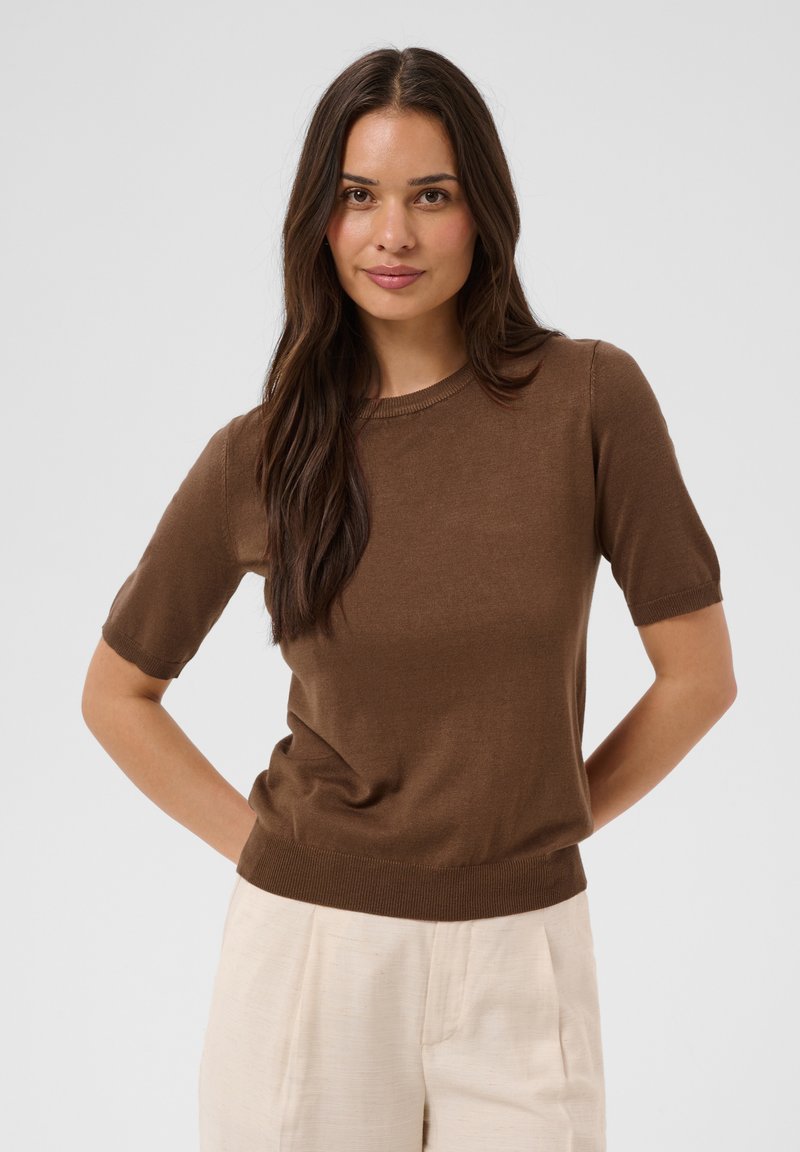 Femme aux longs cheveux foncés portant un haut en maille marron à manches courtes et un pantalon beige clair, debout les mains dans le dos.