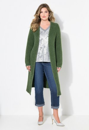 Lange grüne Strickjacke mit offener Front, strukturierter Stoff, silbernes Blumenoberteil, hochgekrempelte Jeans und silberne High Heels.