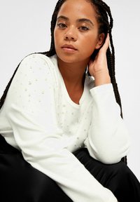 Vit stickad tröja med texturerade, upphöjda pärlliknande detaljer. Långa ärmar och en avslappnad passform. Modellen poserar med flätat hår och minimal sminkning.