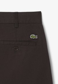 Bruine stoffen broek met een achterzak, knoopsluiting en een klein geborduurd groen krokodil logo. Gladde textuur met een op maat gemaakte stijl.