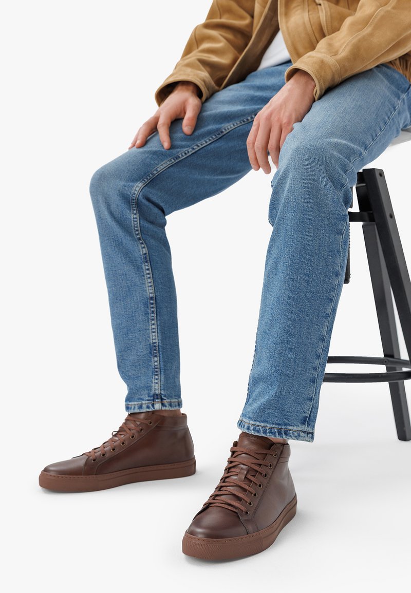 Brown high-top sneakers made of smooth leather, featuring metal eyelets, flat laces, and a patterned rubber sole. Worn with blue jeans.