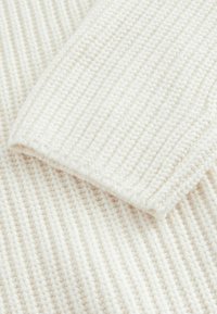 Weißer Strickstoff mit geripptem Muster, sichtbaren Stichen und einem weichen, voluminösen Aussehen. Das Material wirkt dick und gemütlich.