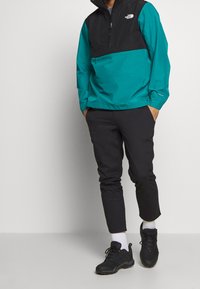 Giacca zip-up teal e nera con maniche lunghe e colletto alto, abbinata a pantaloni neri e sneakers scure. Polsini corti sui pantaloni.