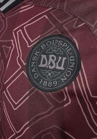 Maglietta sportiva color bordeaux con un pattern testurizzato, un patch rotondo nero con la scritta bianca "DBU" e dettagli intricati nel tessuto.