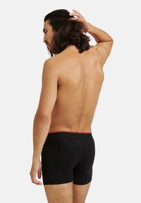 Svarta bomullsboxershorts med en åtsittande design, featuring en röd midjeband. Bakifrån visas en slät, böjd siluett.