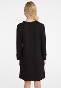 Frau mit blonden Haaren, die ein schwarzes knielanges Langarmkleid trägt, von hinten vor einem weißen Hintergrund gezeigt.