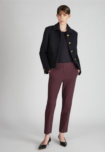 Chaqueta negra de mezcla de lana con botones dorados, combinada con pantalones ajustados burdeos y tacones negros de punta, modelo contra un fondo gris.