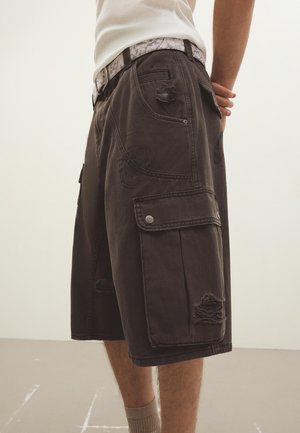 Mand iført mørkegrå, revnede cargoshorts, hvidt mønstret bælte og hvid ærmeløs skjorte, står med hænderne bag ryggen.