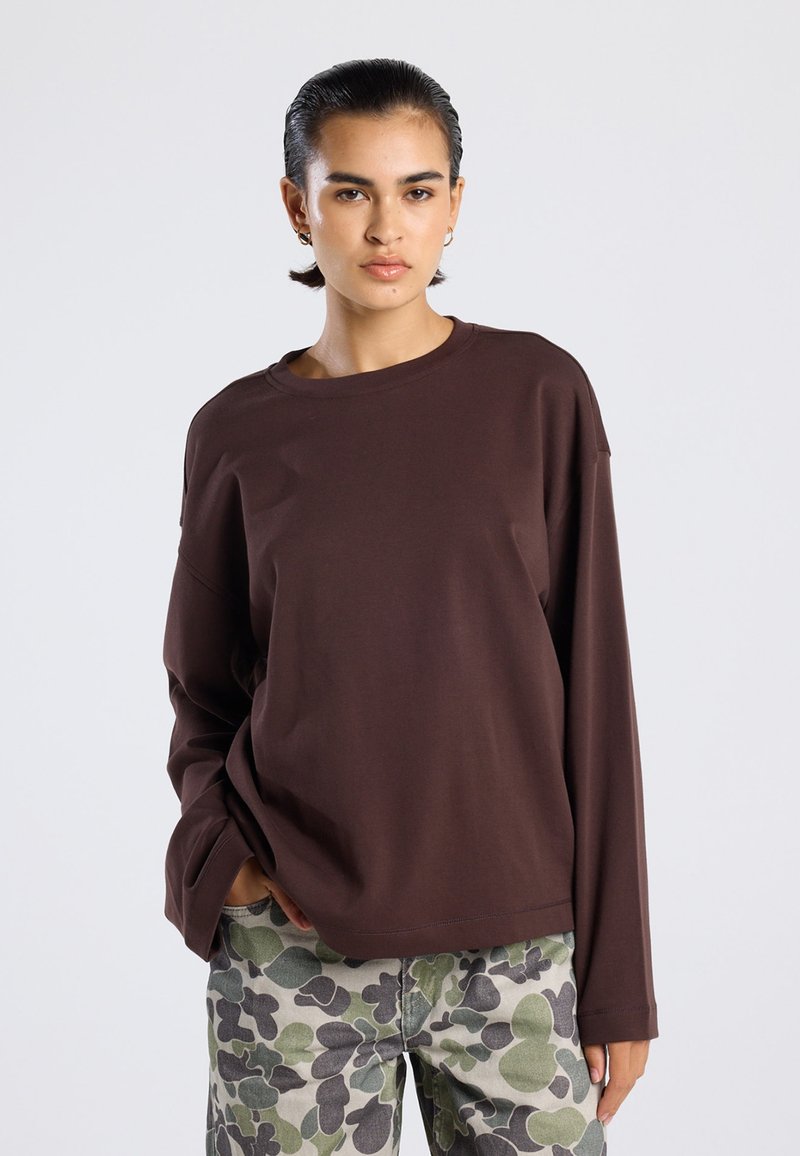 Braunes Langarmshirt aus weichem Material mit Rundhalsausschnitt und lockerer Passform, kombiniert mit gemusterten grünen und grauen Hosen mit Blattmotiven.