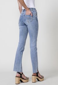 Un jean évasé bleu clair avec une taille haute, des poches arrière et un ourlet brut avec des fentes sur le côté, associé à des sandales à plateforme noires et blanches.