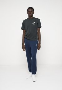 Μαύρη βαμβακερή μπλούζα με μικρή γραφική παράσταση στο αριστερό στήθος, συνδυασμένη με ναυτικά κολάν που διαθέτουν κεντημένο λογότυπο, φορεμένα με λευκά αθλητικά παπούτσια.