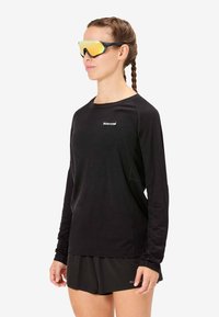 Zwart sportshirt met lange mouwen en een ronde halslijn met logodetail. Gecombineerd met zwarte shorts, model draagt gespiegelde zonnebril met gele tint.