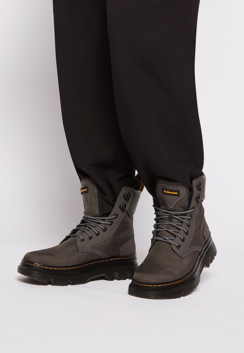 Pantalon noir porté avec des bottes en cuir grises montantes, dotées de semelles épaisses, de lacets noirs et blancs, et d'accents de couture jaune.