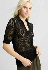 Polo top nero lavorato a maglia con un design in pizzo e motivi geometrici, maniche corte e orlo e colletto a coste nero solido.