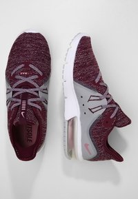 Les baskets Nike Air Max présentent une tige en mesh marron rougeâtre et gris, une semelle blanche, un amorti à air visible au talon, avec des lacets et des détails gris.