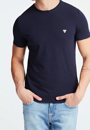 Mężczyzna ubrany w gładki granatowy t-shirt z krótkim rękawem i jasnoniebieskie dżinsy, ręce częściowo w kieszeniach, z małym trójkątnym logo na piersi.