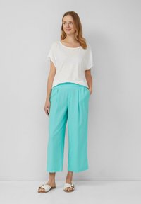 Top bianco a maniche corte e pantaloni larghi color turchese chiaro con tasche. Modella che indossa sandali bianchi con dettaglio a fibbia.