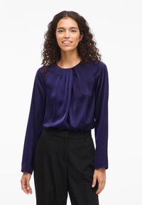 Kvinna med lockigt hår klädd i en lila långärmad blus och svarta byxor, stående mot en ren vit bakgrund, med ett svagt leende.