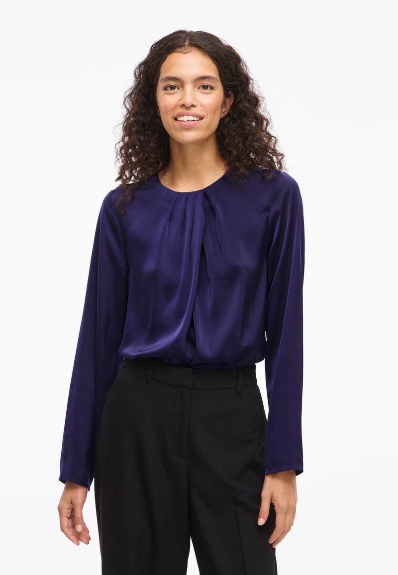 Kvinna med lockigt hår klädd i en lila långärmad blus och svarta byxor, stående mot en ren vit bakgrund, med ett svagt leende.