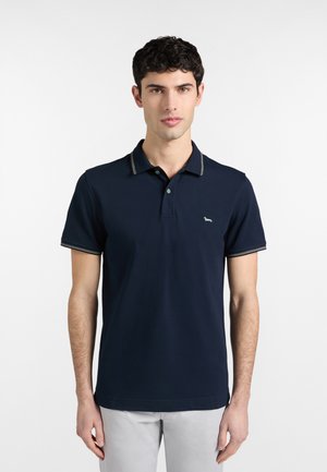 Giovane uomo che indossa una polo blu navy con colletto e maniche a righe, in piedi davanti a uno sfondo bianco semplice.
