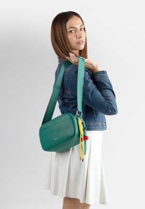 Donna con giacca di denim e gonna bianca che tiene una borsa a tracolla verde con nappe colorate sulla spalla, guardando di lato.