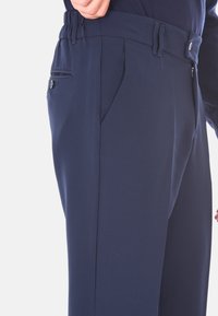 Uomo che indossa pantaloni blu navy su misura con tasche laterali e posteriori, dettaglio dei bottoni e una camicia blu navy infilata dentro.