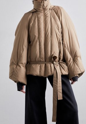 Beige oversized pufferjack met hoge kraag, brede mouwen en een lange riem die om de taille is gestrikt, gedragen over zwarte broek met wijde pijpen.