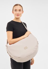Borsa a spalla beige realizzata in materiale morbido, con una forma curva e sovradimensionata e una discreta etichetta del logo. Tracolla regolabile per maggiore comodità.