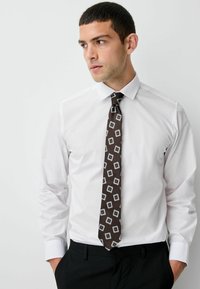 White dress shirt with long sleeves, paired with a brown tie featuring a geometric pattern in gray and white. Black trousers.