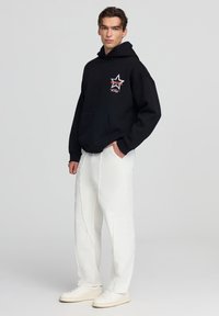 Felpa nera con cappuccio e una grafica a stella bianca sul petto, abbinata a pantaloni bianchi strappati e sneakers bianche.