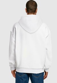 Fehér, túlnyomórészt oversized kapucnis pulóver, nagy, kerek kapucnival és bordázott mandzsettákkal. Puha tapintású, laza fazonú, és látható varrási részletek a szegélyén.