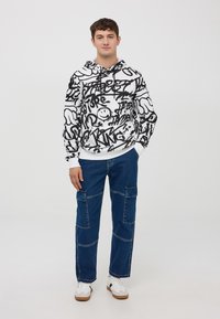 Felpa con cappuccio bianca con motivi di graffiti neri, abbinata a pantaloni cargo blu con molteplici tasche e sneaker bianche.
