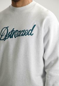 Felpa grigia chiara con scollatura rotonda. Presenta sul davanti la scritta ricamata in teal scuro "Dstressed". Tessuto morbido, vestibilità comoda.
