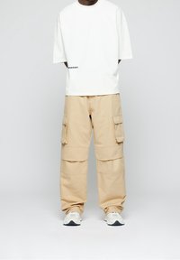 T-shirt blanc oversize avec texte noir sur la gauche. Pantalon cargo beige avec plusieurs poches latérales. Le mannequin porte des baskets grises et blanches.