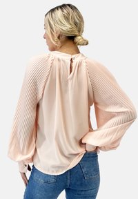 Blusa de melocotón claro con mangas largas con pliegues, escote trasero con forma de ojo de cerradura y dobladillo redondeado. Fabricada en un tejido suave y ligero con una textura lisa.