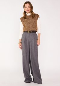 Top senza maniche a maglia marrone su un dolcevita bianco, abbinato a pantaloni grigi a gamba larga con vita alta e una cintura marrone. Sfondo semplice.