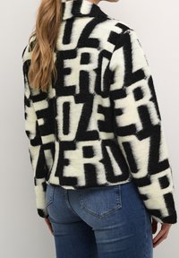 Svartvit fluffig jacka med stora blockbokstäver, krage och avslappnad passform. Kombinerad med blå jeans, vilket visar en avslappnad outfit.