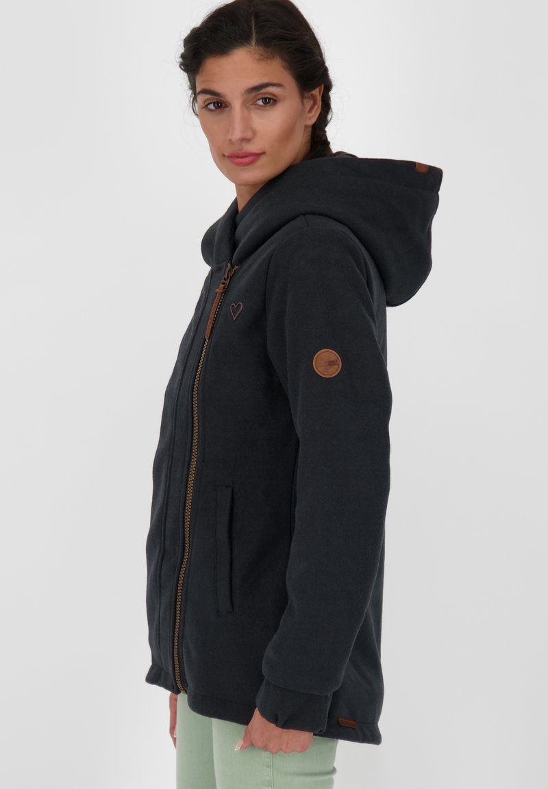 Ciemnoszara kurtka z kapturem, zapinana na zamek, z kieszeniami i brązowymi skórzanymi detalami. Posiada emblemat serca na piersi oraz ściągacze przy mankietach.