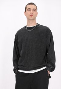Schwarzer, langärmeliger Pullover mit strukturiertem Finish und überschnittenen Schultern, über einem weißen Hemd getragen, kombiniert mit einer schwarzen Hose und einer silbernen Kette.
