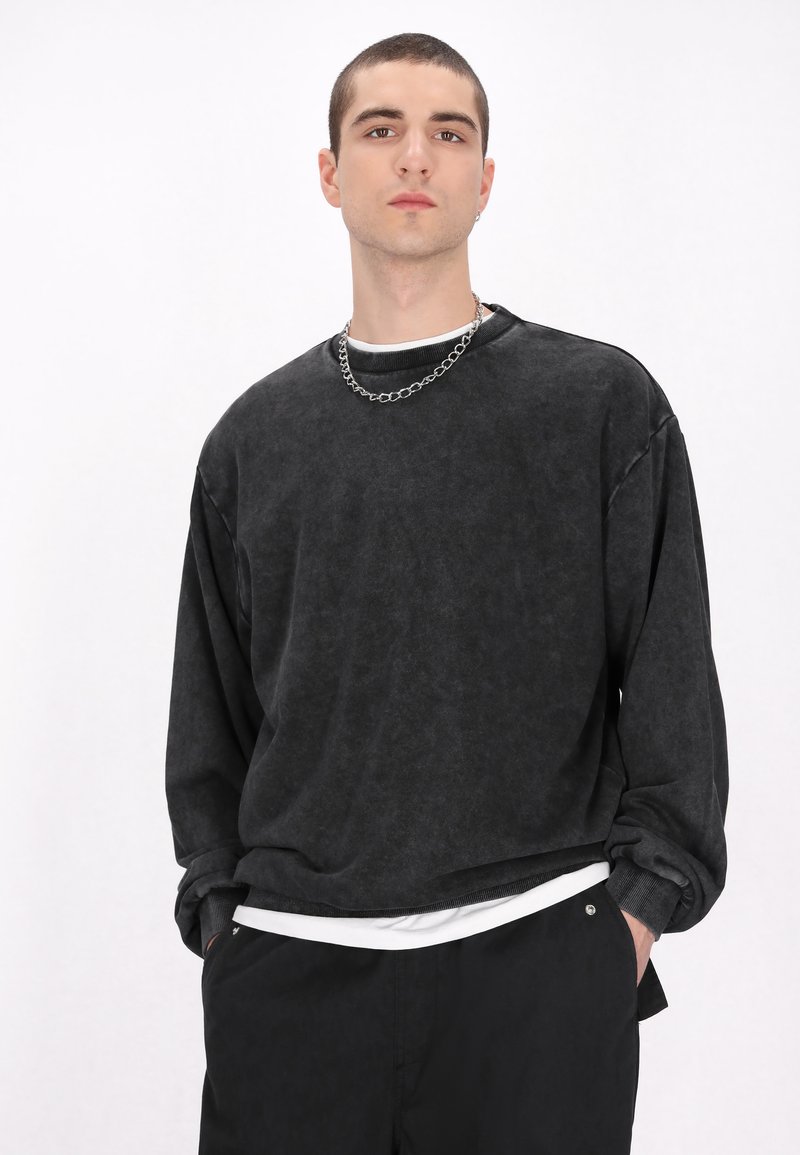 Schwarzer, langärmeliger Pullover mit strukturiertem Finish und überschnittenen Schultern, über einem weißen Hemd getragen, kombiniert mit einer schwarzen Hose und einer silbernen Kette.