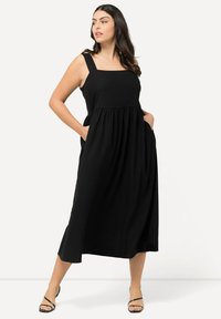 Mustas maxi-mekko, jossa on neliökaula-aukko, paksut hihnat, poimutettu vyötärö ja sivutaskut, valmistettu kevyestä kankaasta.