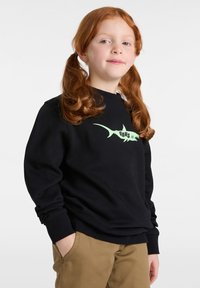 Černá mikina s dlouhými rukávy má zelenou grafiku žraloka a text "VANS". Model má na sobě khaki kalhoty a ruce má v kapsách.