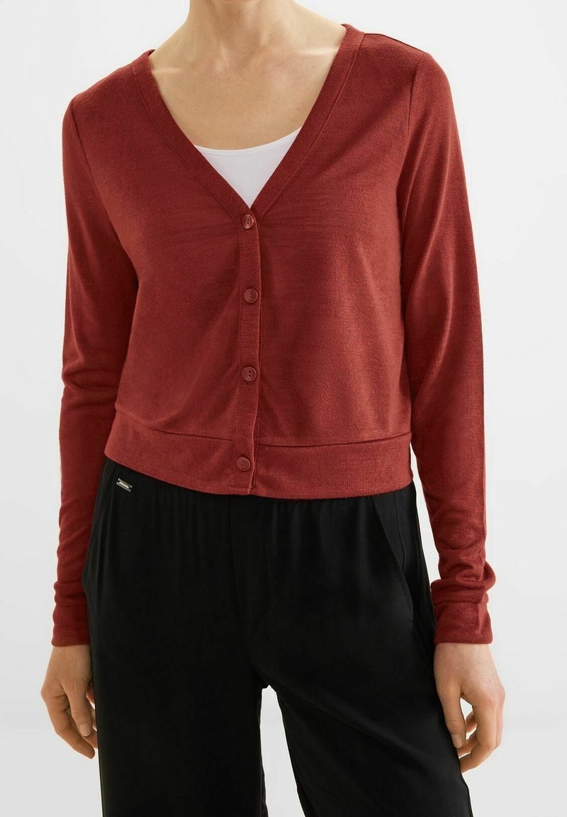 Femme portant un gilet boutonné de couleur rouille sur un haut blanc, associé à un pantalon noir à taille haute, debout devant un fond neutre.