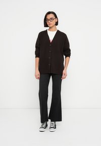 Rjavi rebrasti kardigan z V-izrezom in šestimi gumbi, kombiniran s črnimi širokimi hlačami in črnimi visokimi športnimi copati. Bel ozadje.