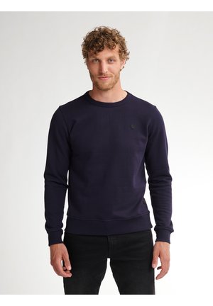 Man met krullend haar, gekleed in een nauwsluitende donkerblauwe sweater met ronde hals en zwarte broek, staand tegen een effen lichte achtergrond.