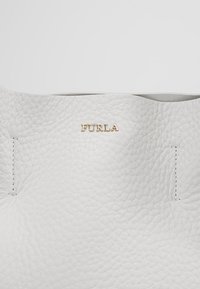 Surface en cuir blanc texturé avec le logo Furla doré centré près du haut et des coutures visibles des deux côtés.