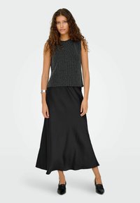 Czarny top bez rękawów z fakturowanym wzorem, zestawiony z długą czarną spódnicą z satyny. Modelka ma na sobie czarne mule i srebrną bransoletkę na nadgarstku.
